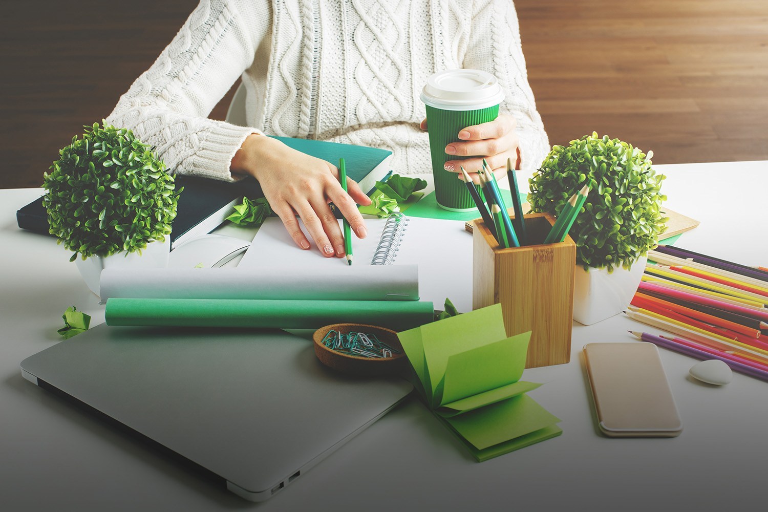 Girl hodling green coffee cup and writing in spiral notepad placed on creative desktop with various items